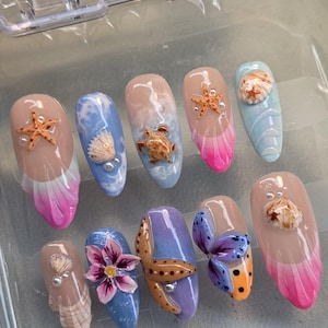 Könnte beinhalten: Ein Set aus zehn künstlichen Fingernägeln mit ozeanischen Motiven, darunter 3D-Seesterne, Muscheln, eine Meeresschildkröte und eine Blume. Die Nägel zeigen verschiedene Farbverläufe in Pink, Blau und Lila mit glänzenden Oberflächen und Perlen.