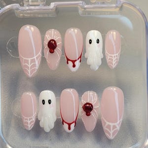 Könnte beinhalten: Ein Set aus zehn ovalen Kunstnägeln in einer transparenten Hülle. Die Nägel haben eine hellrosa Basis mit Designs wie weißen Spinnweben, roten Blutstropfen und weißen Geisterfiguren mit schwarzen Augen. Eine rote Spinne mit einem Edelstein ist ebenfalls abgebildet.