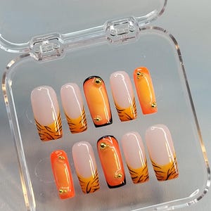 Könnte beinhalten: Ein Set mit zehn falschen Nägeln mit Tigerstreifen-Design. Die Nägel sind hellrosa mit schwarzer Spitze und orangefarbenem Tigerstreifen-Design. Jeder Nagel hat einen goldenen Akzent.
