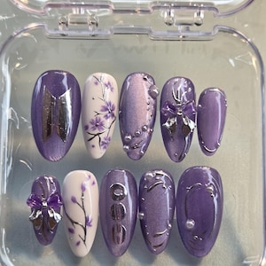 Può includere: Un set di dieci unghie finte a forma di mandorla in una custodia di plastica trasparente. Le unghie presentano una palette di colori viola e bianco con accenti in argento metallizzato, tra cui un logo BTS, motivi floreali e piccole perle.