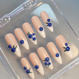 Könnte beinhalten: Set aus zehn mandelförmigen Kunstnägeln. Jeder Nagel hat eine hellrosa Basis mit einer weißen Spitze, verziert mit blauen Blaubeer-Designs und grünen Blättern. Die Nägel werden in einer transparenten Kunststoffbox präsentiert.