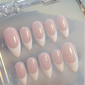 Könnte beinhalten: Ein Set mit zehn falschen Nägeln in einem durchsichtigen Plastikbehälter. Die Nägel sind hellrosa mit weißen Spitzen, was einen klassischen French-Maniküre-Look ergibt.