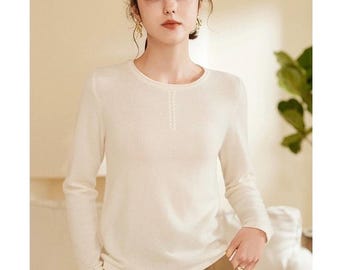 May include: A white long-sleeved sweater with a subtle cable knit detail on the front. The sweater has a round neckline and a relaxed fit.