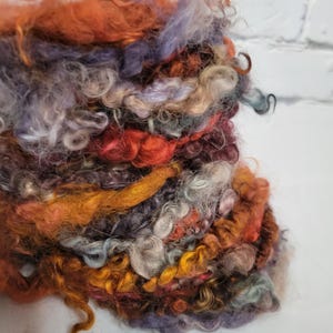 May include: Close-up of a bundle of colorful, textured yarn. The yarn features a mix of orange, purple, yellow, and gray fibers, with a variety of textures including curly and straight strands. The yarn is likely for crafting or textile projects.