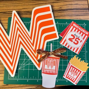 Puede incluir: Una colecci&oacute;n de recortes de papel con tem&aacute;tica de Whataburger, que incluye el ic&oacute;nico logotipo naranja y blanco, un vaso de bebida, un recipiente de papas fritas y un letrero "WHATASENIOR class of '25". Un lazo brillante est&aacute; unido al recorte de la bebida. Los art&iacute;culos est&aacute;n sobre una esterilla de corte verde.