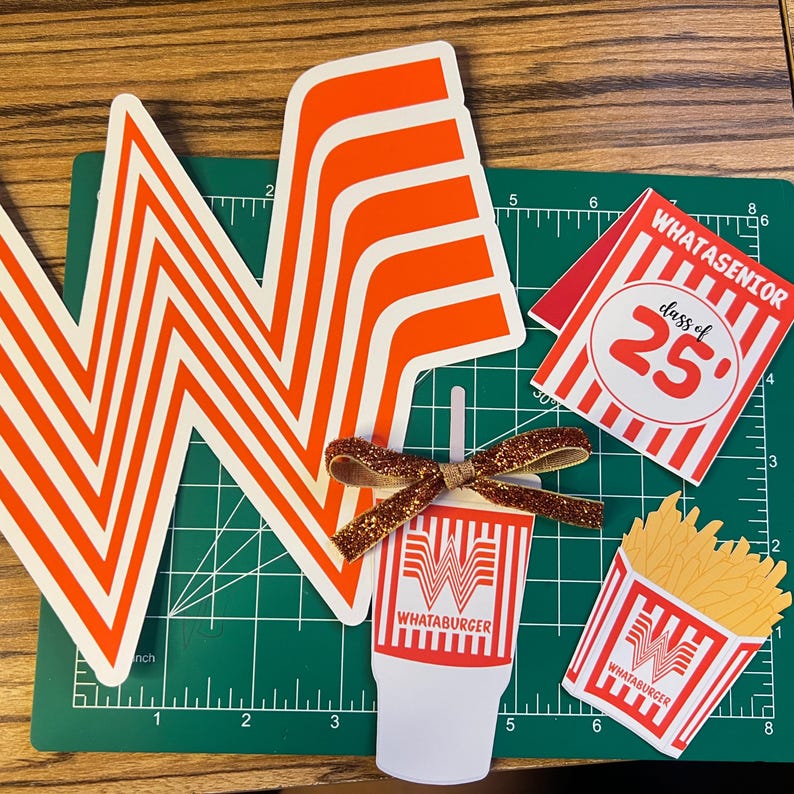 Puede incluir: Una colecci&oacute;n de recortes de papel con tem&aacute;tica de Whataburger, que incluye el ic&oacute;nico logotipo naranja y blanco, un vaso de bebida, un recipiente de papas fritas y un letrero "WHATASENIOR class of '25". Un lazo brillante est&aacute; unido al recorte de la bebida. Los art&iacute;culos est&aacute;n sobre una esterilla de corte verde.