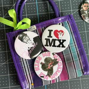 May include: A purple woven tote bag with a lime green ribbon, featuring three circular pin-back buttons. One button shows a man in a baseball cap with the text "LATINAS FOR YANKEE," another says "I LOVE MX" with a heart, and the third says "KISS ME I'M MEXICAN."