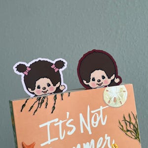May include: Two die-cut paper bookmarks featuring cartoon characters with brown hair, pink bows, and rosy cheeks, positioned at the top edge of a peach-colored book cover with the text It's Not Summer and illustrations of a sand dollar and seaweed.