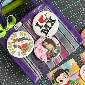 May include: A small purple woven bag with a green ribbon, featuring three circular pin-back buttons. One button says I LOVE MX with a heart, another says KISS ME I'M MEXICAN with a couple dancing, and the third says baila mi corazón with a person.
