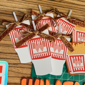 Puede incluir: Una colecci&oacute;n de recortes de papel con tem&aacute;tica de Whataburger. Los recortes incluyen vasos blancos con rayas rojas y el logotipo de Whataburger, adornados con lazos y pajitas doradas con purpurina. Tambi&eacute;n se incluyen cajas de patatas fritas.