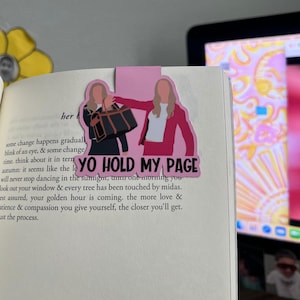 May include: Pink and black bookmark featuring two women in a cartoon style. The women are holding a black purse and wearing pink and red outfits. The text "YO HOLD MY PAGE" is printed on the bookmark.
