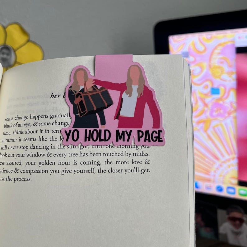 May include: Pink and black bookmark featuring two women in a cartoon style. The women are holding a black purse and wearing pink and red outfits. The text "YO HOLD MY PAGE" is printed on the bookmark.