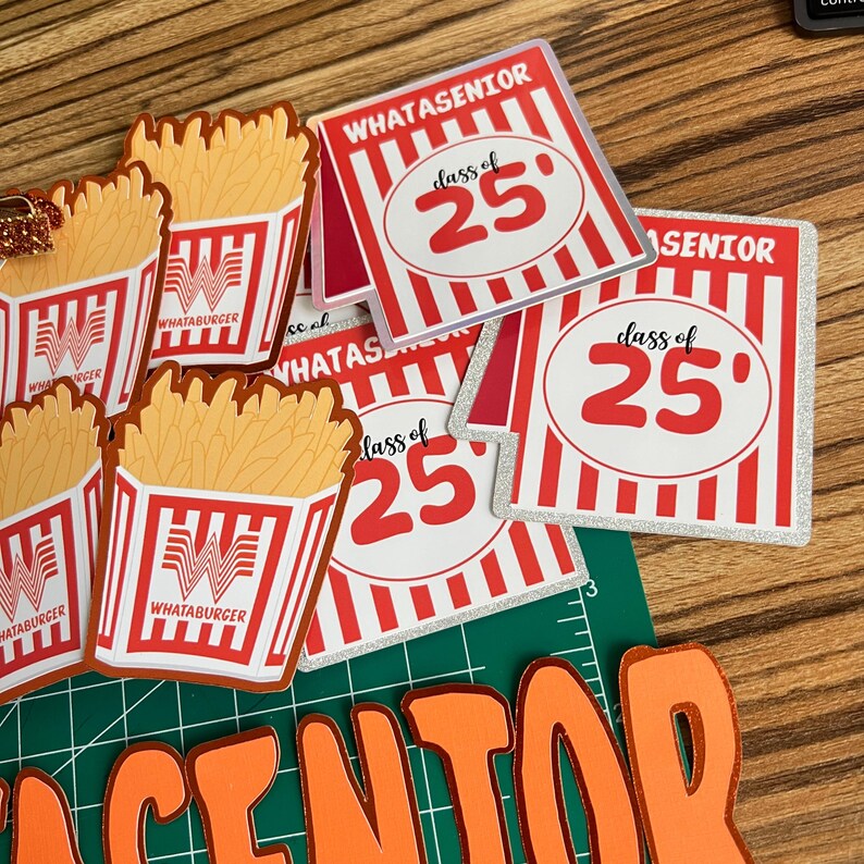 Puede incluir: Una colecci&oacute;n de decoraciones de graduaci&oacute;n con tem&aacute;tica Whataburger. Incluye recortes de papas fritas y tarjetas cuadradas con el texto "WHATASENIOR" y "class of 25" en rojo y blanco. El fondo es una superficie de madera.