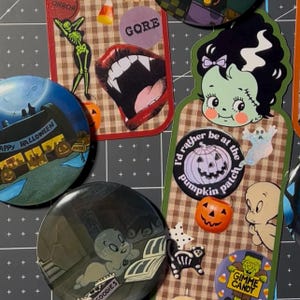 May include: A collection of Halloween-themed items including circular buttons and stickers featuring a green monster, a ghost, a pumpkin with the text 'I'd rather be at the pumpkin patch', a vampire mouth, a skeleton, and a 'Happy Halloween' sign.