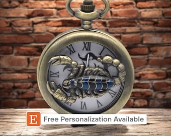 Puede incluir: Un reloj de bolsillo dorado con una esfera de reloj de números romanos y un diseño de escorpión. El escorpión es azul y negro con detalles dorados.