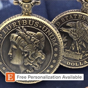 May include: A gold-toned pocket watch with a design of a woman's profile on one side and an eagle on the other. The watch face reads "E. Pluribus Unum" and "United States of America." The back of the watch reads "In God We Trust" and "Dollar."