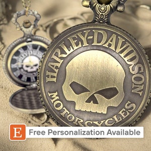 May include: A gold-toned pocket watch with a Harley-Davidson logo and skull design. The watch has a black face with Roman numeral hour markers and a black leather strap.