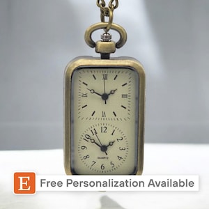 May include: A gold-toned pocket watch with a square face and two separate clock faces. The top clock face has Roman numerals and the bottom clock face has Arabic numerals. The watch is on a chain and has the text "Free Personalization Available" on the bottom.