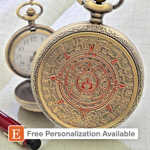 May include: A detailed, antique-style pocket watch with a bronze finish. The watch face features an intricate Aztec calendar design with red accents. A second pocket watch is visible in the background. The image includes the text "Free Personalization Available".