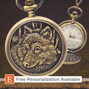 Op de afbeelding: Antieke messing zakhorloge met een gedetailleerd wolfskopontwerp. Het horloge heeft een ketting en een tweede horloge is zichtbaar op de achtergrond. De tekst "Gratis personalisatie beschikbaar" staat onderaan.