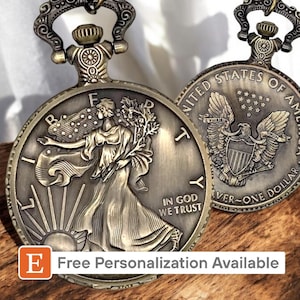 Könnte beinhalten: Eine goldfarbene Taschenuhr mit einem Motiv der Freiheitsstatue auf der einen Seite und einem Adler mit den Worten "United States of America" und "One Dollar" auf der anderen Seite.