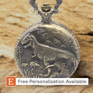 May include: A gold-toned pocket watch featuring an intricate engraving of a dinosaur in a prehistoric scene. The watch has a chain attached to the top.