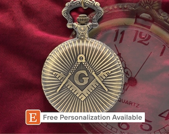 May include: A gold-toned pocket watch with a Masonic symbol on the face. The watch has a  square and compass design with a 'G' in the center. The watch is on a red background.