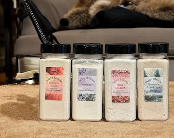 May include: Four glass jars with white powder and black lids. Each jar has a label with a different scent: Pumpkin Spice, Lavender, Rosy Delight, and Season's Greetings. The jars are on a brown fuzzy surface.