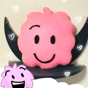 May include: A pink, cloud-shaped plush toy with a black embroidered smiling face and a single raised eyebrow. The plush is positioned in front of a dark crescent moon-shaped shelf. The text "PUFFBALL PLUSH!!!" appears at the bottom of the image.