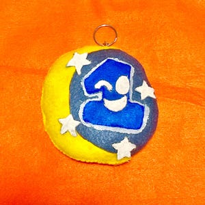 May include: A handmade felt keychain featuring a blue number one with a winking face, set against a gray and yellow moon shape. White star accents surround the design, with a silver keyring at the top. The background is orange.