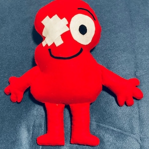 May include: A red plush toy shaped like a figure with a white bandage over one eye and a large white and black eye. It has arms, legs, and a smiling mouth. The toy is made of soft fabric.