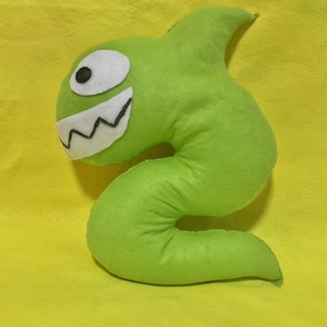 May include: A bright green plush toy in the shape of a worm-like creature. It features a white felt eye with a black pupil and a white felt mouth with jagged teeth. The toy is set against a yellow backdrop.