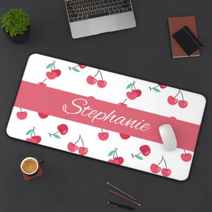 May include: A white rectangular mousepad with a pattern of red cherries and green leaves. A pink band across the center displays the name "Stephanie" in white script. A white computer mouse rests on the right side.
