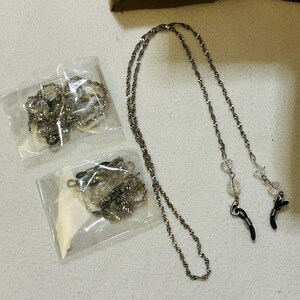 May include: Silver-toned eyeglass chains with clear beads and black rubber loops. Two chains are packaged in clear plastic, and one chain is loose. The background is a light-colored surface.