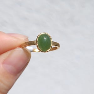 Könnte beinhalten: Ein goldener Ring mit einem glatten, ovalen grünen Stein in einer Fassung. Die Ringschiene hat eine texturierte, matte Oberfläche, die einen Kontrast zur polierten Oberfläche des grünen Edelsteins bildet. Der Ring wird vor einem hellen Hintergrund gehalten.