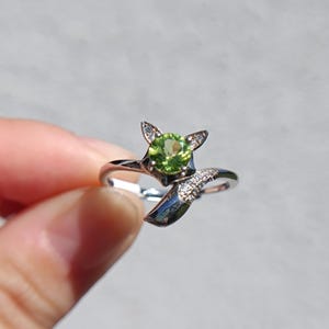 Könnte beinhalten: Ein silberfarbener Ring mit einem fuchsartigen Design, das in der Mitte einen runden, facettierten, lindgrünen Edelstein aufweist. Die Ohren und der Schwanz des Fuchses sind mit kleinen, klaren, funkelnden Steinen besetzt.