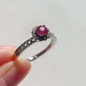 Könnte beinhalten: Ein silberner Ring mit einem runden, tiefroten Edelstein in der Mitte, umgeben von kleineren, klaren Steinen. Das Band hat ein filigranes Design mit kleinen, dunklen Akzenten. Der Ring wird vor einem weißen Hintergrund gehalten.