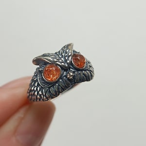 Könnte beinhalten: Ein silberfarbener Ring in Form eines Eulenkopfes mit texturierten Federdetails und zwei runden, durchscheinenden orangefarbenen Steinen als Augen. Das Metall hat ein dunkles, oxidiertes Finish in den Vertiefungen, um die Konturen hervorzuheben.