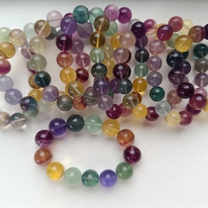 Könnte beinhalten: Eine Sammlung von elastischen Armbändern aus polierten, kugelförmigen Fluoritperlen. Die Perlen haben ein durchscheinendes, mehrfarbiges Aussehen mit natürlichen Bändern in violetten, grünen, gelben und klaren Tönen, die kreisförmig angeordnet sind.