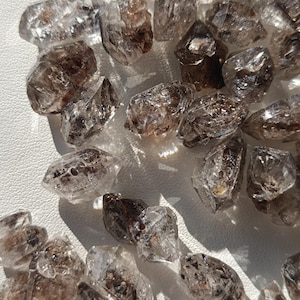 Könnte beinhalten: Eine Ansammlung roher, durchscheinender Quarzkristalle mit natürlichen Einschlüssen und dunkelbraunen bis schwarzen Mineralablagerungen. Jeder Kristall weist unregelmäßige, facettierte Formen mit unterschiedlicher Klarheit auf.