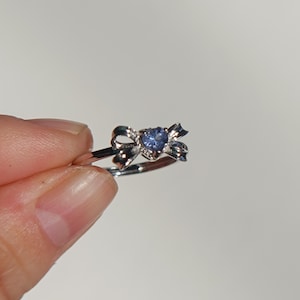 Könnte beinhalten: Ein silberner Ring mit einem Schleifendesign und einem herzförmigen, hellblauen Edelstein in der Mitte. Der Ring wird vor einem schlichten weißen Hintergrund gehalten, um die filigranen Details des Schmucks hervorzuheben.