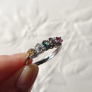 Könnte beinhalten: Ein silberfarbener Ring mit drei rund geschliffenen Edelsteinen in Gelb, Grün und Pink, die jeweils von einem Kranz aus kleinen klaren Steinen umgeben sind. Die Ringschiene ist schmal und metallisch, mit einer blütenförmigen Fassung.
