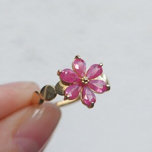 Könnte beinhalten: Ein goldener Ring mit einem blumenförmigen Mittelstück, das aus sechs birnenförmig geschliffenen, leuchtend rosa Edelsteinen besteht, die um eine zentrale Goldperle angeordnet sind. Die Ringschiene hat eine glatte, polierte Oberfläche mit Metallakzenten.