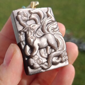 Könnte beinhalten: Rechteckiger Anhänger mit einem detaillierten Relief eines Fuchses inmitten wirbelnder Muster. Der Anhänger hat eine gedämpfte Silberfarbe und ist an einer goldfarbenen Kordel befestigt. Der Fuchs ist der zentrale Fokus des Designs.