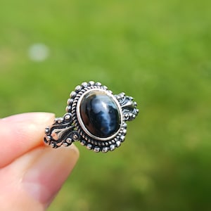 Könnte beinhalten: Ein Silberring mit einem ovalen, dunkelblau-schwarzen Edelstein mit Katzenaugeneffekt, gefasst in einen dekorativen Metallrahmen mit Perlenverzierungen und verzierten Seitenelementen. Die Ringschiene hat ein texturiertes, oxidiertes Finish.