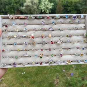 Könnte beinhalten: Eine Präsentation zahlreicher Ringe mit verschiedenen Edelsteinfassungen, darunter Smaragde, Amethyste und Opale. Die Ringe sind in einer weißen Schale angeordnet und zeigen eine Vielzahl von Designs und Farben. Die Ringe sind auf silbernen Bändern gefasst.