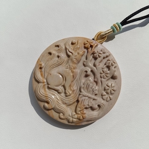 Könnte beinhalten: Ein runder, beigefarbener Steinanhänger mit einer detaillierten Reliefschnitzerei eines Fuchses mit buschigem Schwanz, eines Schmetterlings und floralen Elementen. Der Anhänger hängt an einer schwarzen Kordel mit einem goldenen und hellblauen Knoten.