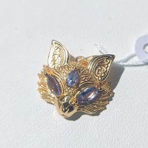 Könnte beinhalten: Ein goldfarbener Metallanhänger in Form eines Fuchskopfes mit komplizierten Gravurmustern an Ohren und Stirn. Der Fuchs hat drei hellviolette, birnenförmige Edelsteine als Augen und Stirnakzent sowie ein kleines weißes Etikett an der Seite.