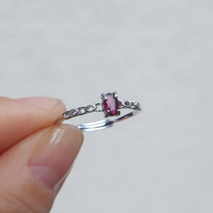 Könnte beinhalten: Ein silberfarbener Ring mit einem kleinen, ovalen, dunkelrosa Edelstein in einer Vier-Krappen-Fassung. Die Ringschiene weist an den Seiten ein zartes, durchbrochenes Muster auf und ist unten offen, um eine flexible Anpassung zu ermöglichen.
