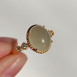 Könnte beinhalten: Ein goldfarbener Ring mit einem großen, ovalen, glatten, hellgrauen Cabochon-Stein in einer perlenartigen Metallfassung. Die Ringschiene ist auf beiden Seiten mit jeweils zwei kleinen, quadratisch gefassten hellblauen Steinen verziert.